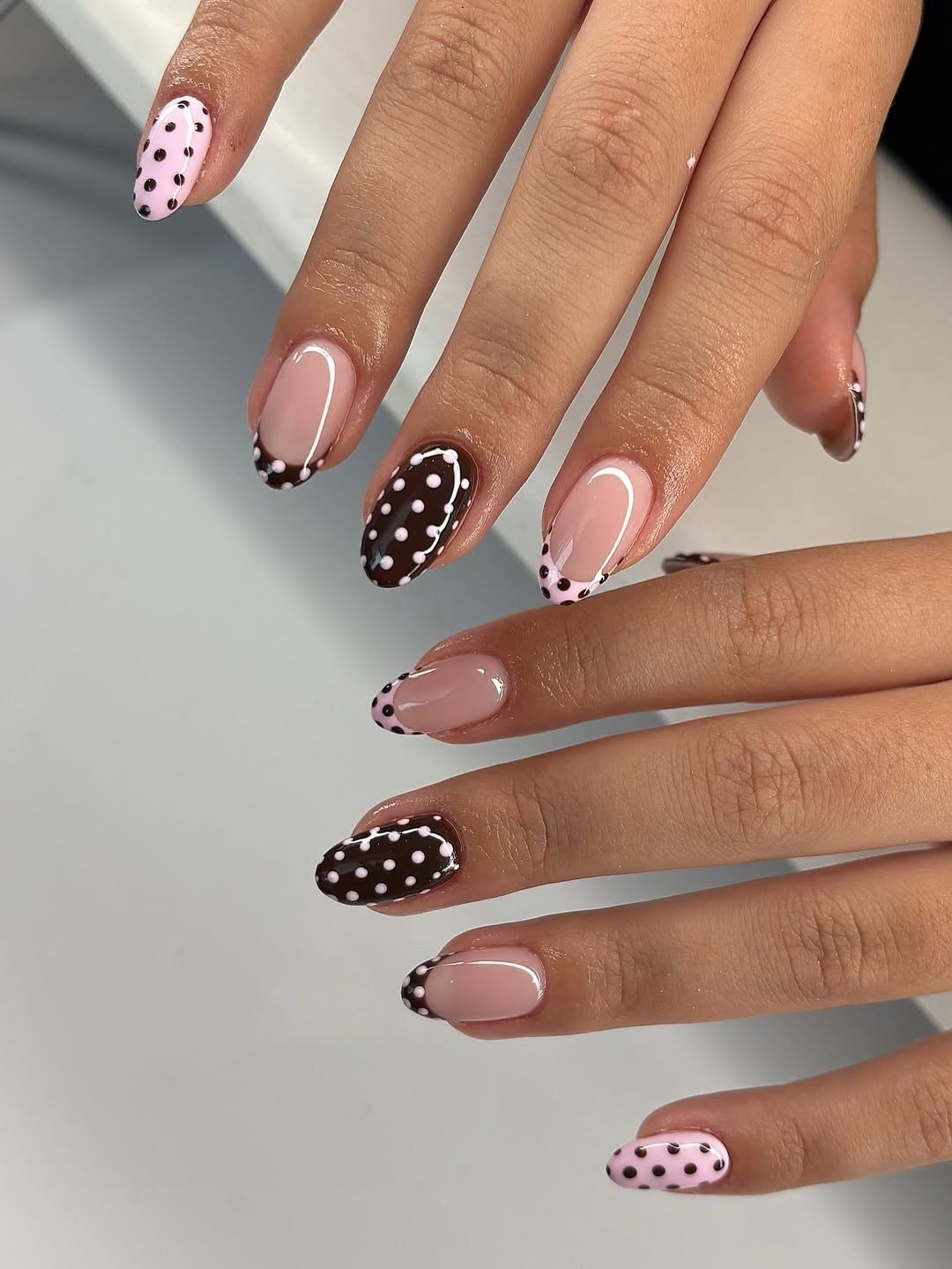 Almond-shaped nails with a mix of light pink with black dots, dark brown with white dots, and French tips with polka dots at…