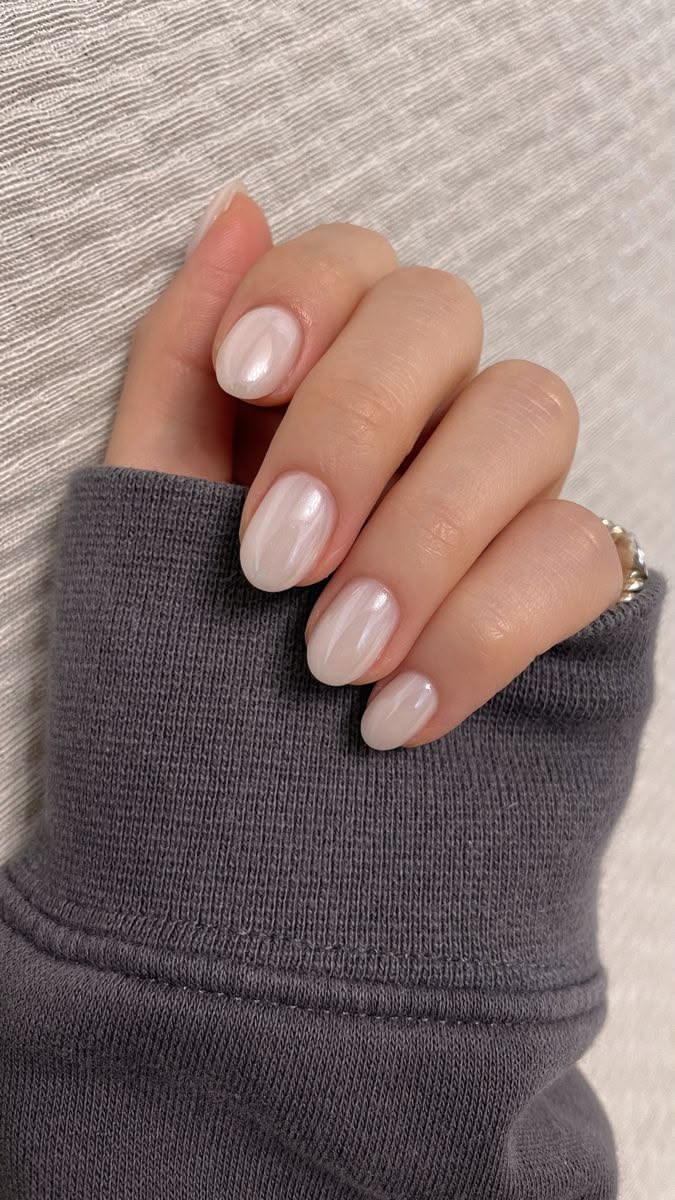 Close-up of a hand with short, oval nails painted with an iridescent, sheer white polish, contrasting with a dark grey…