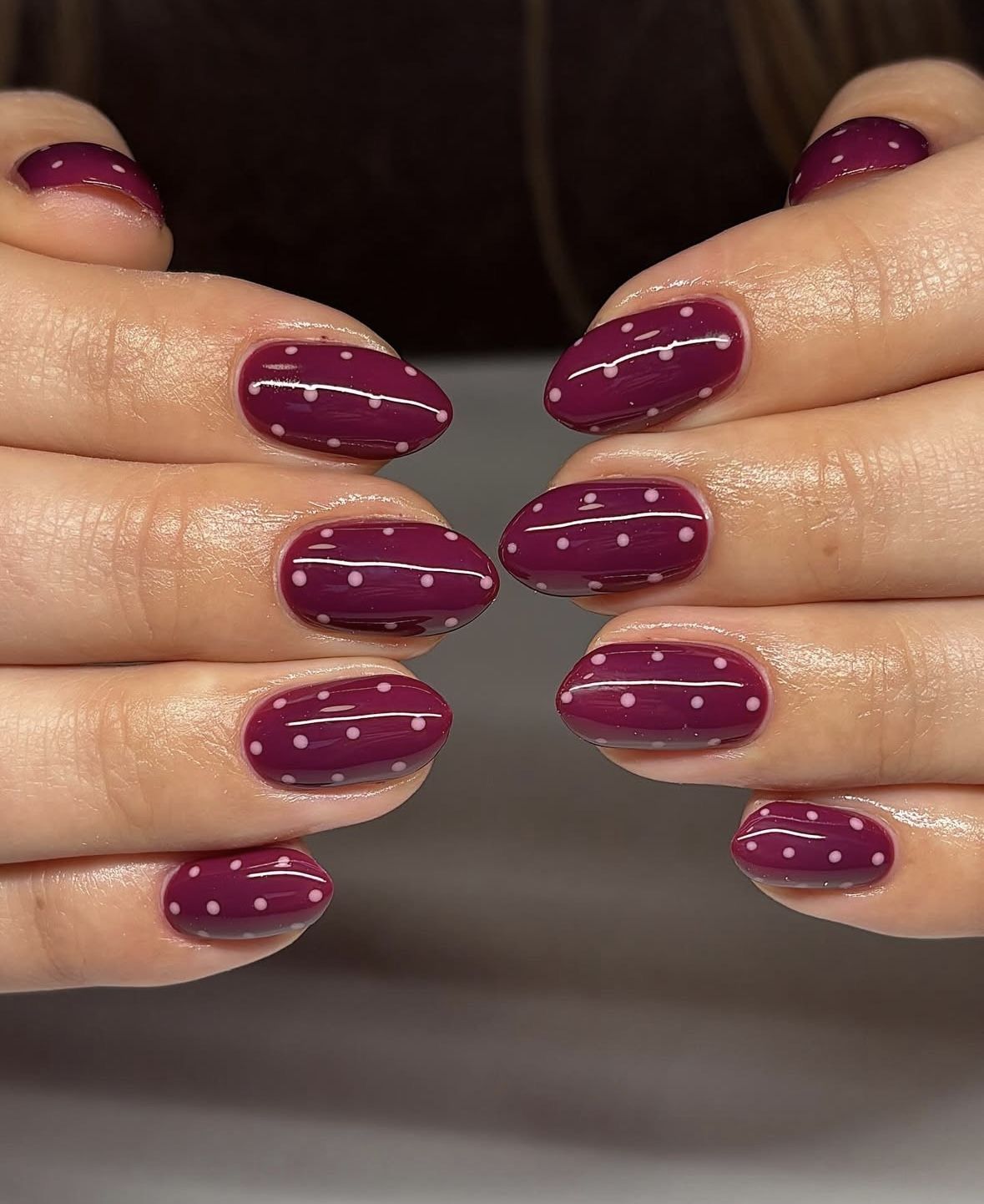 Close-up of hands with burgundy oval nails featuring small white polka dots and thin white horizontal stripes.