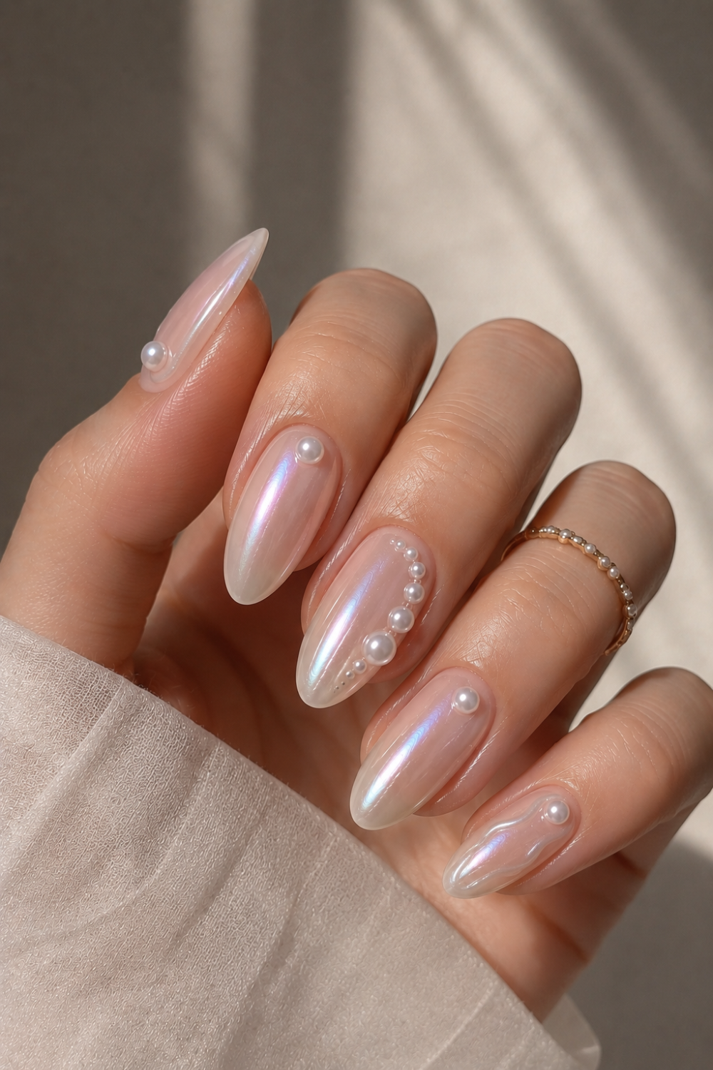 A hand with oval-shaped, iridescent pearl-white nails, some adorned with small 3D pearls and a gold ring on the ring finger.