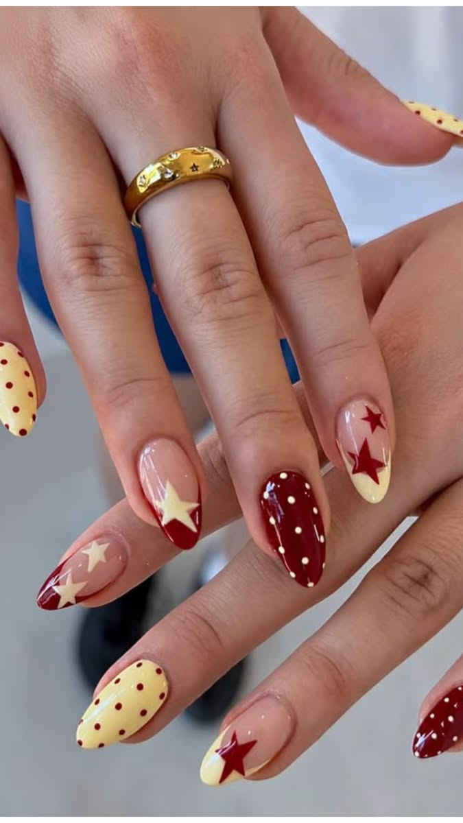 Close-up of manicured hands with almond-shaped nails. Designs feature red and yellow polish with stars and polka dots.