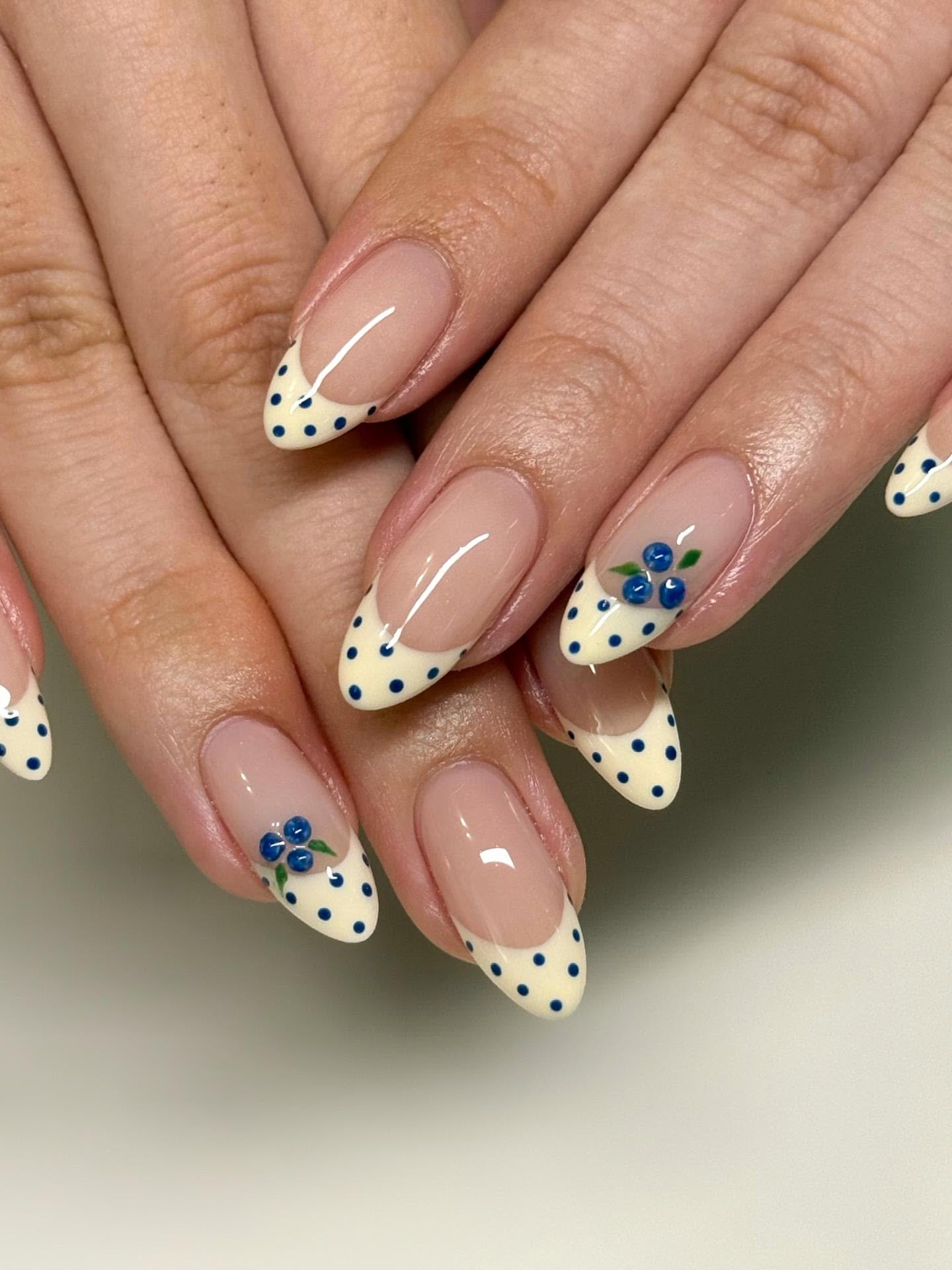 Close-up of hands with manicured nails featuring French tips, yellow polka dots, and blueberry illustrations.