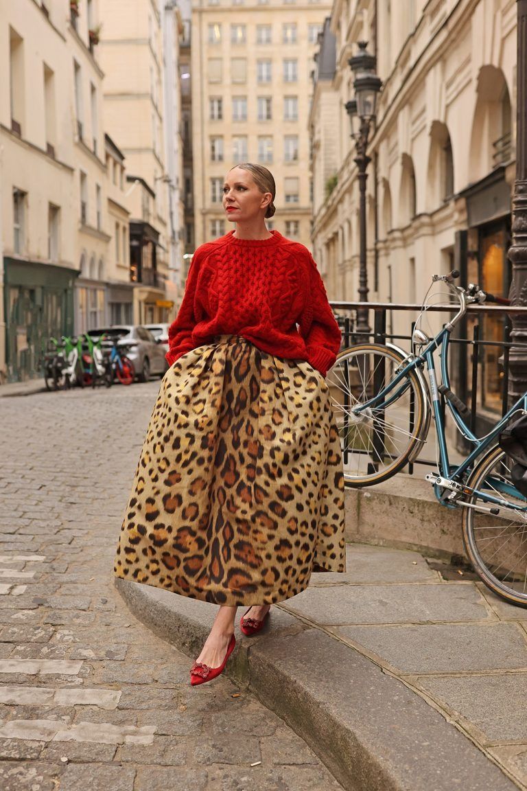 A woman in a vibrant red cable-knit sweater and a voluminous leopard print maxi skirt stands on a cobblestone street.