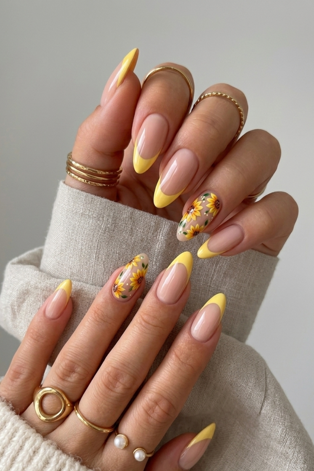 Close-up of hands with long, almond-shaped nails featuring yellow French tips and sunflower art on accent nails, adorned…