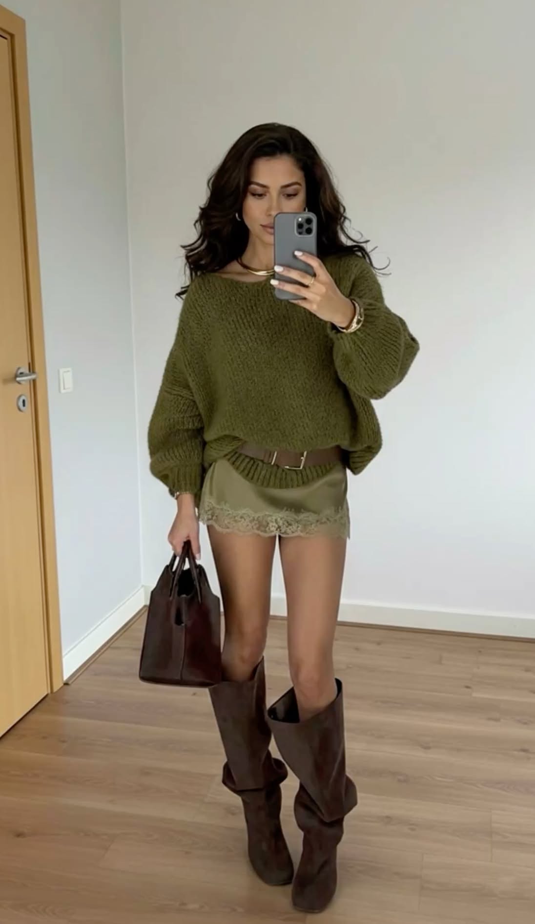 A woman models an olive green oversized sweater over a satin mini-skirt with lace trim, a belt, and brown knee-high boots.