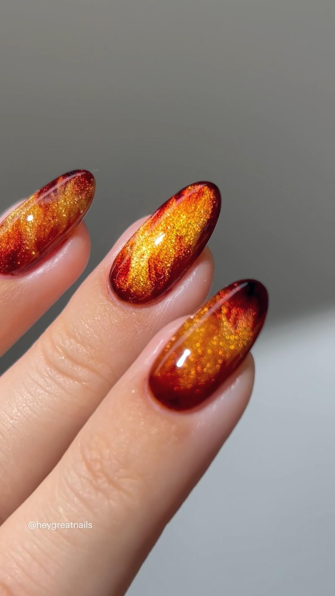 Close-up of three almond-shaped nails with a glossy amber and red marble pattern, shimmering with gold flecks.