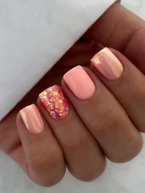 A close-up of a hand with square-tipped nails. Most nails are painted a solid light coral, with two shimmering metallic…