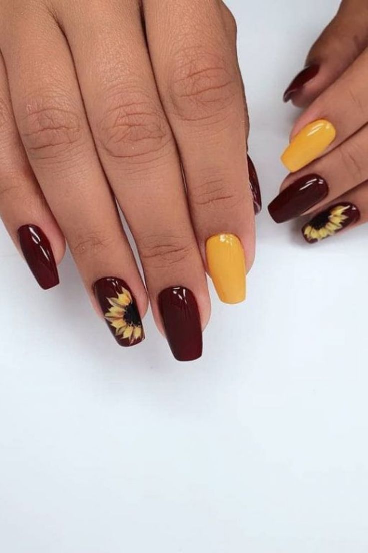 Manicured hand with coffin-shaped nails. Alternating nails are painted deep reddish-brown and mustard yellow, with two nails…