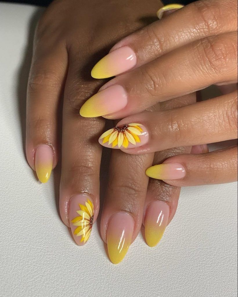 Close-up of manicured hands with almond-shaped nails featuring a yellow to nude ombre gradient and two nails with sunflower…