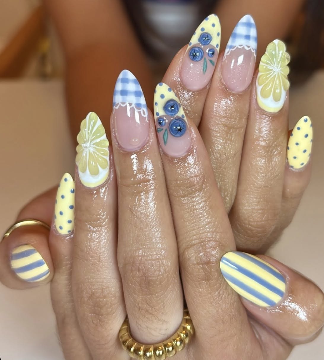 Close-up of hands with varied almond-shaped nail art including blue gingham tips, blueberries, lemon slices, yellow polka dot