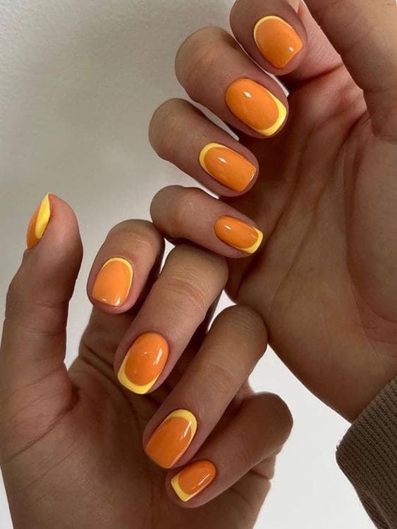 Close-up of hands with short, oval nails painted bright orange with yellow French tips