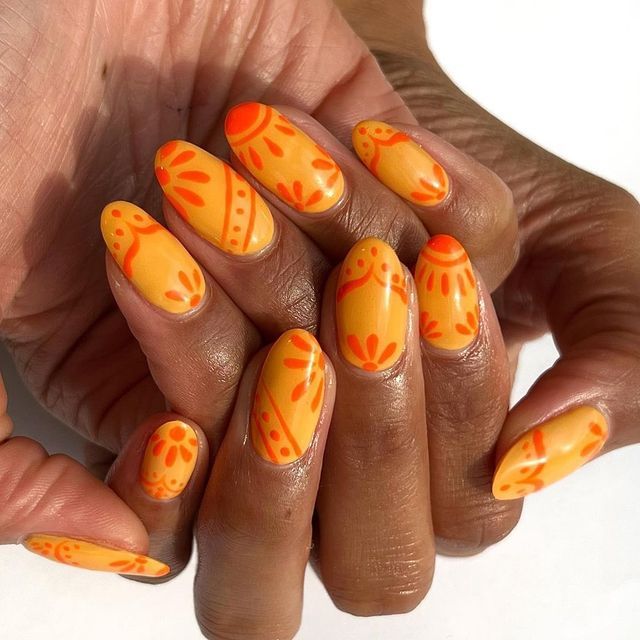 Close-up of a hand with yellow oval nails adorned with orange mandala and dots patterns.