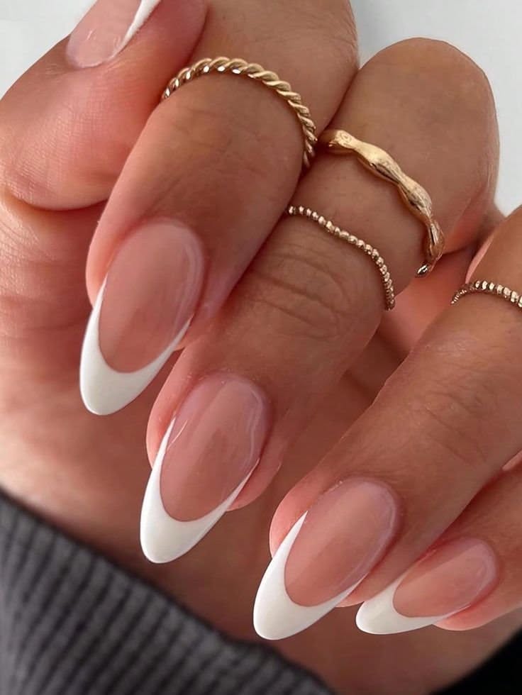 Close-up of a hand with almond-shaped white French tip nails and multiple thin gold rings on the fingers.