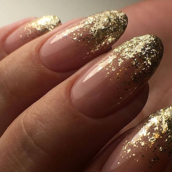 Close-up of almond-shaped nails with a blush pink base and gold glitter flakes fading from the tips.