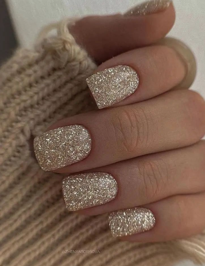 Close-up of a hand with short, square nails painted in dense, warm gold glitter resting on a beige knit sweater.
