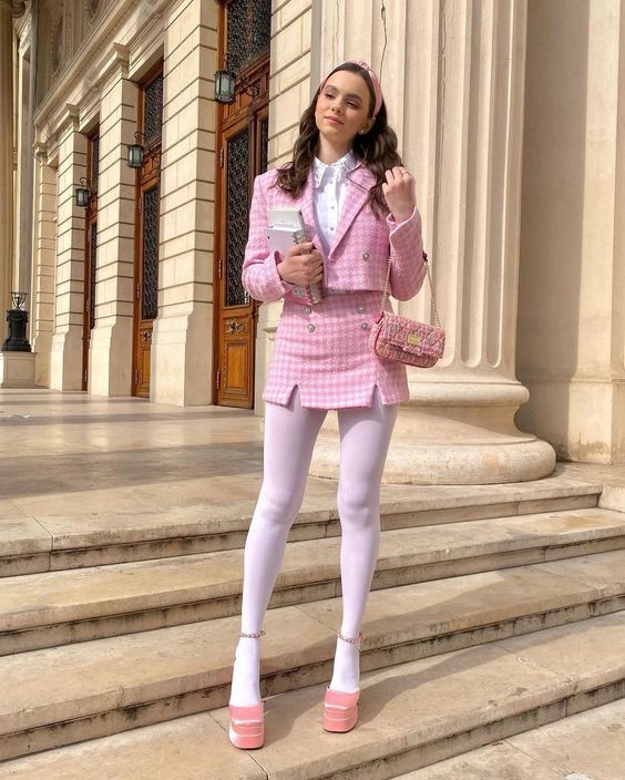 A woman wears a pink tweed cropped jacket and matching mini skirt, white button-down shirt, white tights, and pink platform h